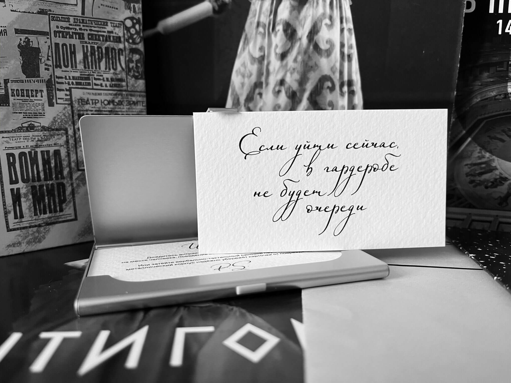 Карточка «Если уйти сейчас, в гардеробе не будет очереди» стоит в алюминиевой визитнице на фоне театральных афиш